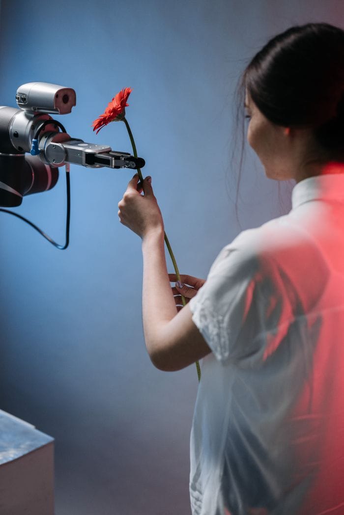services-01 A robotic arm delicately holds a red flower as a woman interacts with it.
